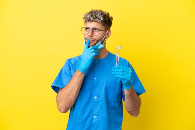 Dentysta Kaukaski Mężczyzna Trzymający Narzędzia Na Białym Tle Na żółtym Tle Mający Wątpliwości Podczas Patrzenia W Górę