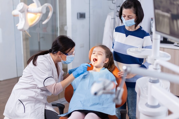 Dentysta korzystający z lustra w trakcie badania jamy ustnej dziecka siedzącego na fotelu dentystycznym. Lekarz stomatolog podczas konsultacji jamy ustnej dziecka w gabinecie stomatologicznym z wykorzystaniem nowoczesnych technologii.