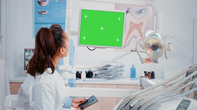 Dentysta patrząc na monitor z poziomym zielonym ekranem do pielęgnacji jamy ustnej w gabinecie stomatologicznym. Kobieta pracująca jako stomatolog z szablonem makiety i izolowanym tłem do pielęgnacji zębów