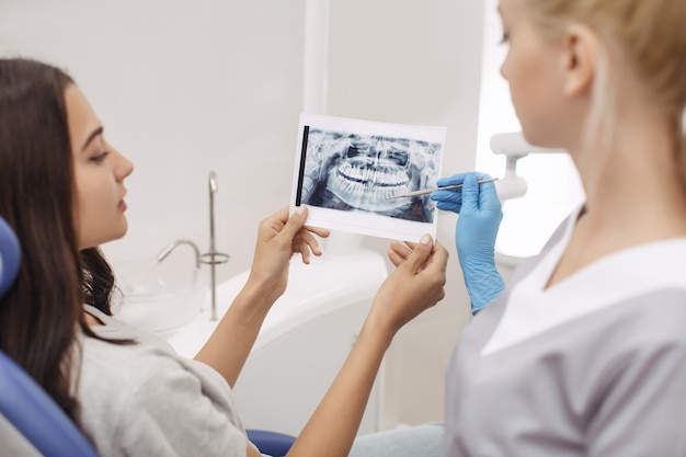 Dentysta pokazujący pacjentowi zdjęcie rentgenowskie. Ludzie, medycyna, stomatologia, technologia i koncepcja opieki zdrowotnej - szczęśliwa kobieta dentysta z obrazem rentgenowskim zębów i pacjentką w biurze kliniki dentystycznej