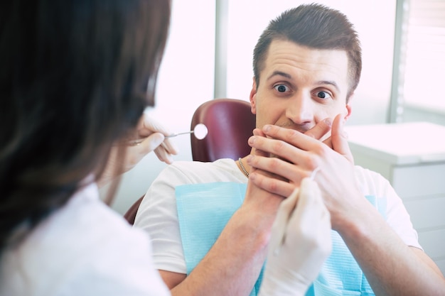 Dentysta Próbuje Leczyć Mężczyznę, Ale Nie Może, Ponieważ Jest Bardzo Przestraszony I Pokazuje To Z Rękami Zakrytymi Ustami I Spojrzeniem Na Instrumenty