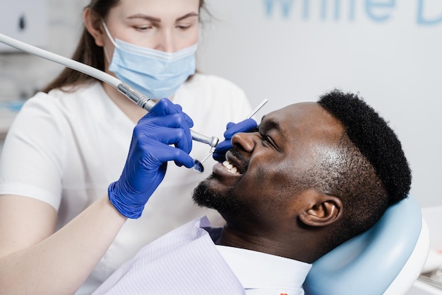 Dentysta z wiertłem dentystycznym leczy afrykańskiego mężczyznę w klinice dentystycznej Wypełnienie dentystyczne dla pacjenta afroamerykanina