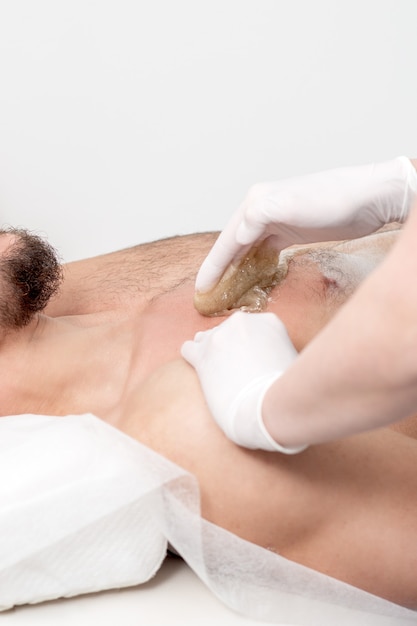 Depilacja klatki piersiowej młodego mężczyzny z płynną pastą woskową rękami kosmetyczki w gabinecie kosmetycznym