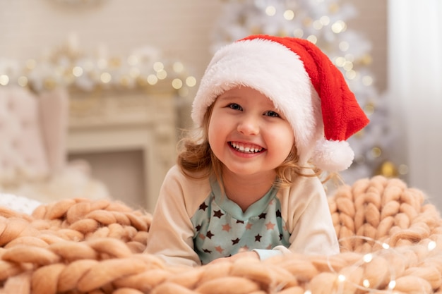 Designerskie Koce Z Beżu Merino! śmiejące Się Dziecko W Santa Hat. Za Dziewczyną W Tle Jest Choinka Przy Oknie