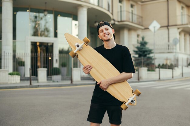 deskorolkarz facet pozuje z longboardem w dłoniach na ulicy miasta, patrząc na kamerę