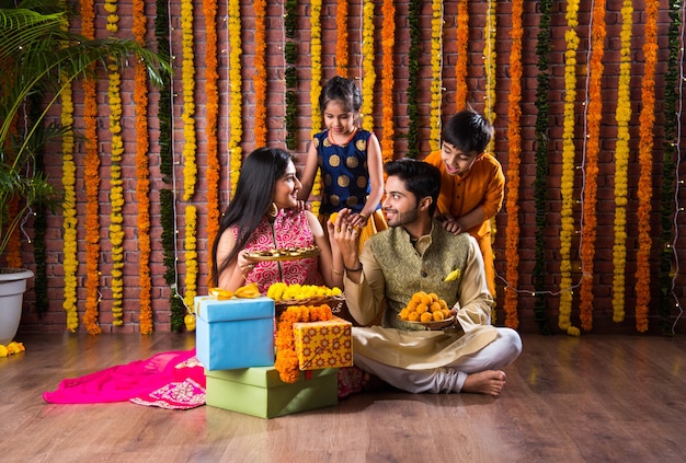 Diwali Lub Rakshabandhan Celebration - Indyjska Młoda Czteroosobowa Rodzina świętuje Festiwal Deepavali Lub Bhai Dooj Ze Słodkim Laddoo, Lampą Oliwną Lub Diya I Pudełkami Prezentowymi, Jedząc Jedzenie Lub Robiąc Selfie