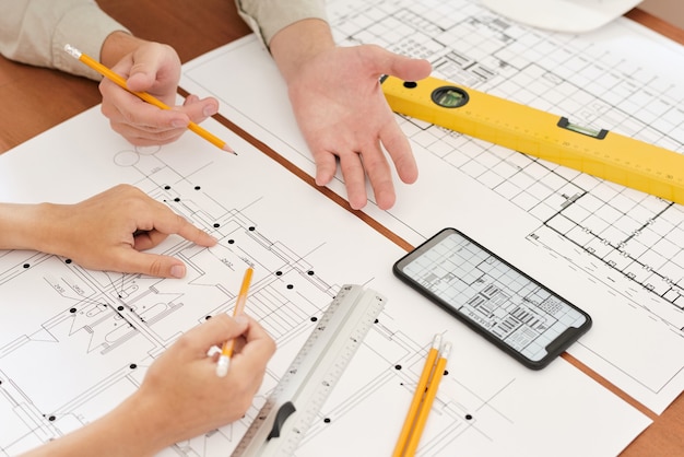 Dłonie dwóch młodych inżynierów z ołówkami nad szkicami nowej konstrukcji podczas omawiania jej szczegółów i kreatywnych pomysłów