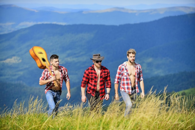 Długa trasa. Grupa młodych ludzi w kraciastych koszulach chodzących razem na szczycie góry. Mężczyźni z gitarą w słoneczny dzień. Koncepcja turystyki pieszej turystów. Piesze wycieczki z przyjaciółmi. Wspólna radość z wolności.