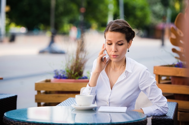Dobra rozmowa biznesowa. Rozochocony młody bizneswoman opowiada na telefonie komórkowym i trzyma filiżankę kawy podczas gdy siedzący outdoors