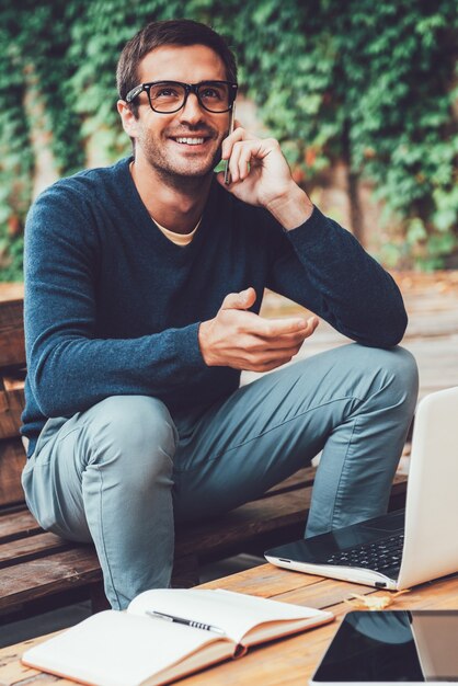 Dobra rozmowa biznesowa. Szczęśliwy młody człowiek rozmawia przez telefon komórkowysiedząc przy drewnianym stole na zewnątrz