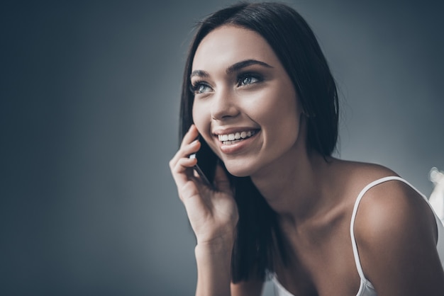 Dobra rozmowa z przyjacielem. Atrakcyjna młoda kobieta rozmawia przez telefon komórkowy hte i uśmiecha się siedząc przy szarej ścianie