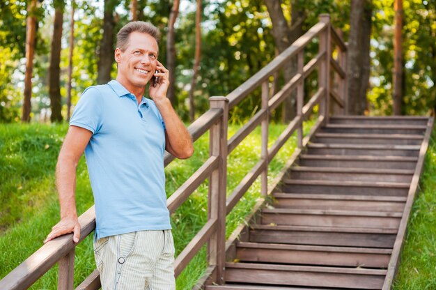 Dobra rozmowa z przyjacielem. Wesoły dojrzały mężczyzna rozmawia przez telefon komórkowy i uśmiecha się stojąc przy drewnianych schodach