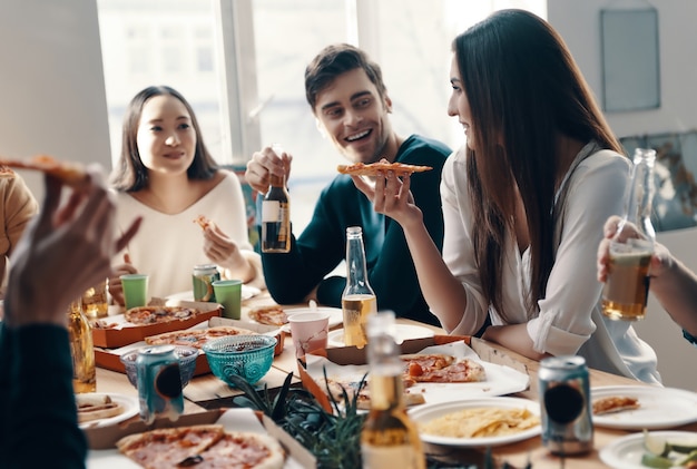 Dobre jedzenie i dobrzy przyjaciele. Grupa młodych ludzi w strojach codziennych jedzących pizzę i uśmiechających się podczas kolacji w pomieszczeniu