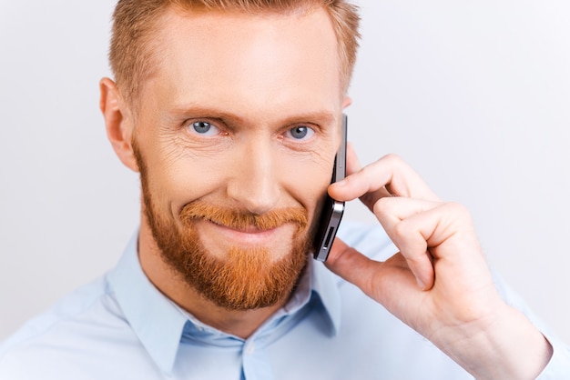 Dobre wieści. Zbliżenie: brodaty mężczyzna rozmawia przez telefon komórkowy i uśmiecha się stojąc na białym tle