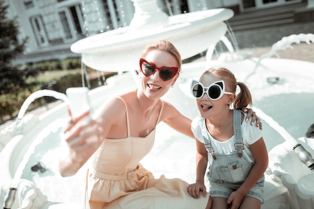 Dobrze się bawić. Wesoła matka i córka siedzi przy pięknej fontannie i robi śmieszne miny biorąc selfie.