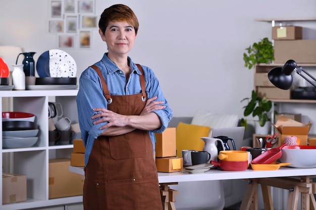 Zdjęcie dojrzała azjatycka kobieta przedsiębiorca / właścicielka firmy stojąca przed swoim glinianym produktem ceramicznym i pracująca w domu
