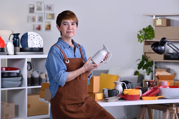 Dojrzała azjatycka kobieta przedsiębiorca / właścicielka firmy stojąca przed swoim glinianym produktem ceramicznym i pracująca w domu