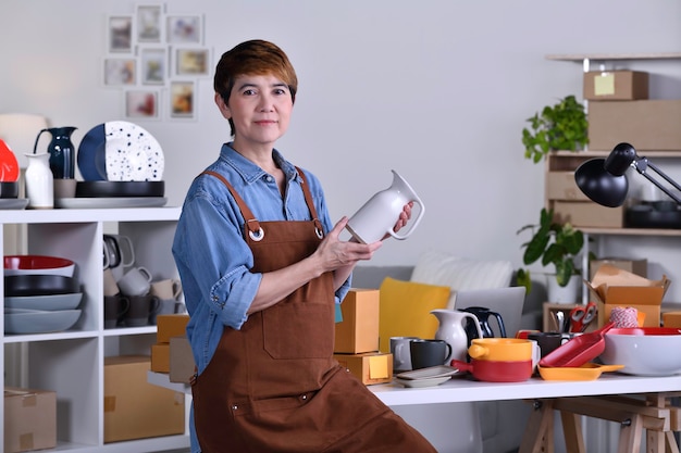 Zdjęcie dojrzała azjatycka kobieta przedsiębiorca / właścicielka firmy stojąca przed swoim glinianym produktem ceramicznym i pracująca w domu