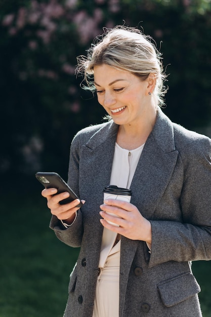 Dojrzała blond uśmiechnięta kobieta biznesu spacerująca po parku, pijąca kawę i rozmawiająca przez telefon na wiosnę na tle kwiatów bzu