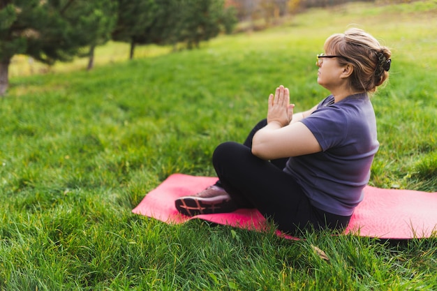 Dojrzała emerytka rasy kaukaskiej w parku medytuje i wykonuje ćwiczenia oddechowe w letni dzień