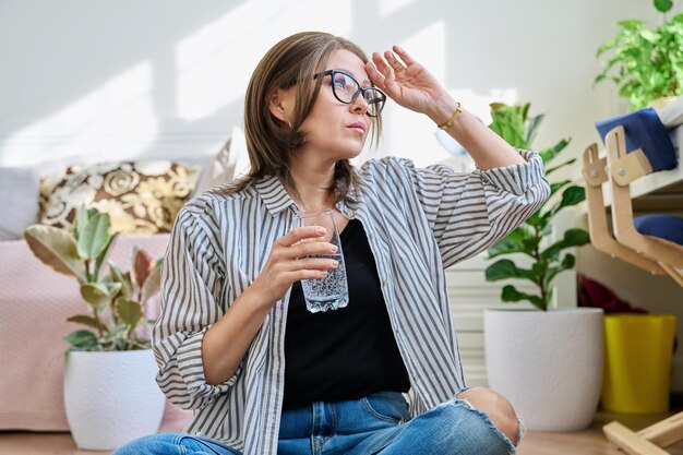 Dojrzała kobieta cierpiąca na ból głowy w domu ze szklanką wody