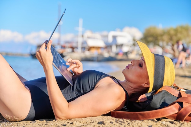 Dojrzała kobieta opalająca się na plaży w kapeluszu z laptopem