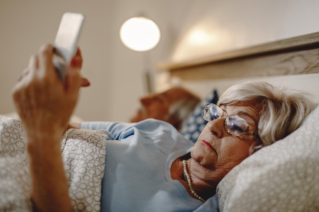 Dojrzała kobieta relaksuje się w łóżku i czyta SMS-a na telefonie komórkowym Jej mąż śpi za nią