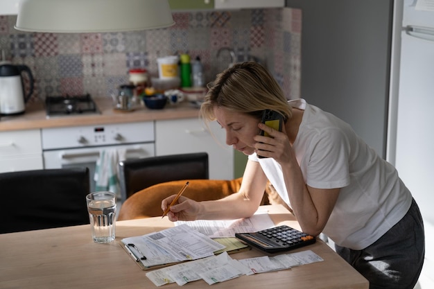 Dojrzała kobieta rozmawia przez telefon z infolinią, próbując rozwiązać problem sprawdzania niezapłaconych rachunków przy stole