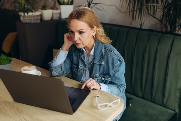 Dojrzała kobieta siedzi w kawiarni przed laptopem i myśli o problemie, dotykając głowy rękami Koncepcja freelance