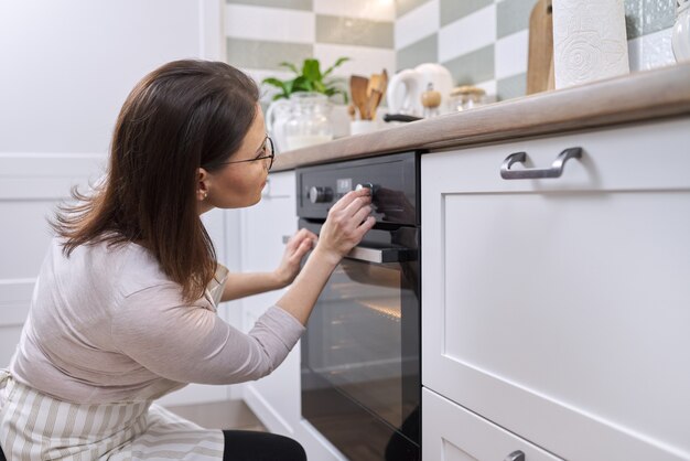 Dojrzała kobieta w fartuchu w pobliżu piekarnika w kuchni. Gospodyni włącza ustawienia na kuchence, kopiuje przestrzeń