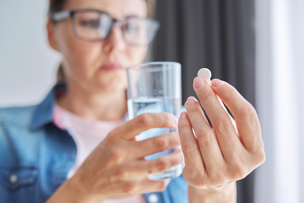 Dojrzała kobieta w średnim wieku w codziennych ubraniach w domu trzymająca pigułkę i szklankę świeżej wody Bóle głowy depresja leki suplementy witaminy środki uspokajające menopauza