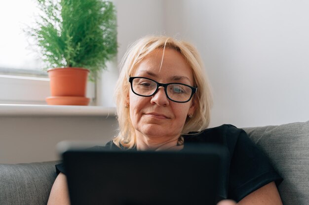 Dojrzała kobieta w średnim wieku zajmująca się e-bookami w domu na kanapie