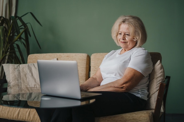 Dojrzała kobieta za pomocą laptopa przeglądania Internetu siedzi na kanapie, pracując zdalnie, patrząc na ekran w domu Koncepcja starości i ludzi technologii