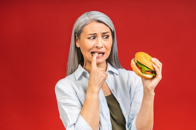 Dojrzała starsza kobieta je burgera z satysfakcją Babcia lubi smaczny hamburger na wynos pyszny kęs burgera zamów dostawę fastfood, gdy głodny stoi na białym tle na czerwonym tle