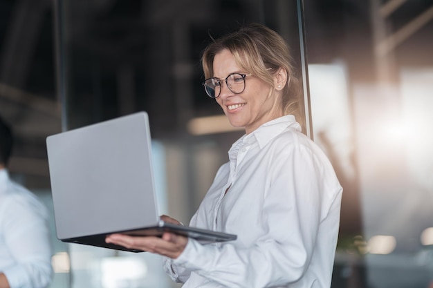 Dojrzała uśmiechnięta bizneswoman w nowoczesnym biurze pracuje na laptopie