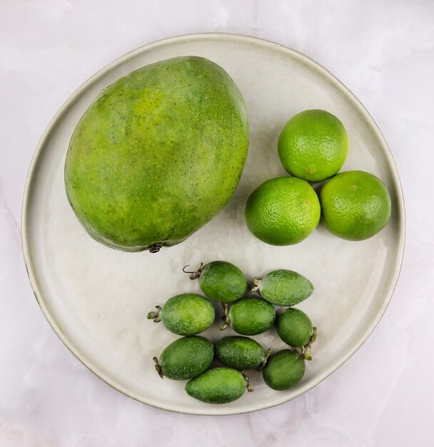 Dojrzałe owoce mango całe jagody feijoa i całe cytryna na talerzu izolowanym na szarym marmurowym tle Zielony trend spożywczy Egzotyczne superfoody zdrowa koncepcja jedzenia