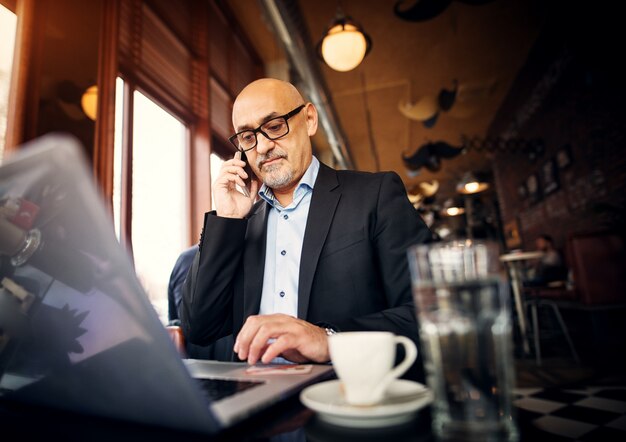 Dojrzały Fachowy Biznesmen Siedzi Przy Stołem Sklep Z Kawą Robi Rozmowie Telefonicza I Używa Laptop