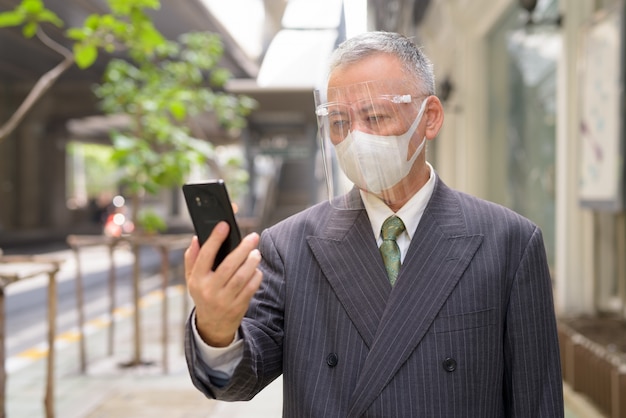 Dojrzały Japoński biznesmen z maską i twarzy osłoną używać telefon w mieście