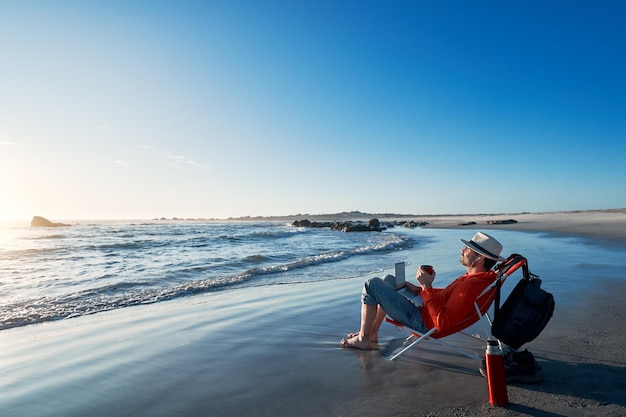 Dojrzały Latynos siedzi na brzegu plaży z laptopem pijąc kawę i ciesząc się