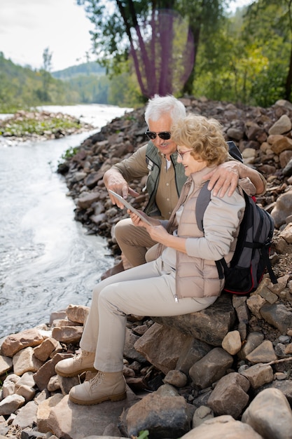 Dojrzały mężczyzna i jego żona z plecakami patrzą na ekran touchpada, siedząc na brzegu rzeki nad wodą