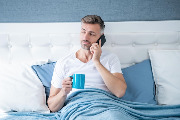 Dojrzały mężczyzna pije kawę w łóżku i rozmawia przez telefon