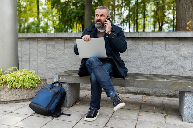 Dojrzały mężczyzna pracujący na laptopie na zewnątrz rozmawiający przez telefon