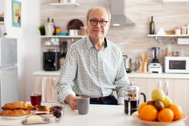 Dojrzały mężczyzna ze zmarszczkami patrząc na kamery w nowoczesnej kuchni przy filiżance kawy. Portret zrelaksowanej starszej osoby starszej rano, pijącej świeży ciepły napój, zdrowa uśmiechnięta twarz dorosłego