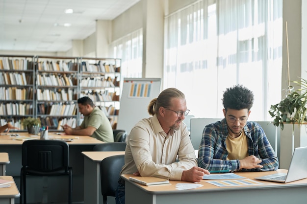 Dojrzały nauczyciel uczy języka obcego studenta, siedząc przy biurku w bibliotece