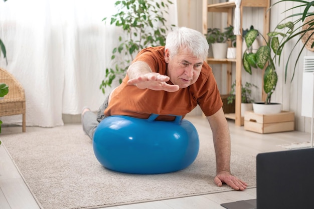 Dojrzały starszy mężczyzna robi ćwiczenia fitness gimnastyka w domu Lekcja online z laptopem Rozmowa trenera Rekreacja dobre samopoczucie Starszy mężczyzna ćwiczący trening rozciągający Stary człowiek ćwiczący