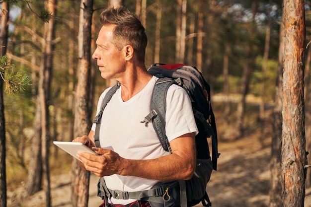 Dojrzały, wysportowany mężczyzna z plecakiem spaceruje po lesie i korzysta z internetu na nowoczesnym touchpadzie