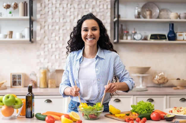 Dokonywanie zdrowego posiłku szczęśliwa latynoska kobieta gotuje świeżą sałatkę, stojąc w kuchni i uśmiechając się do kamery