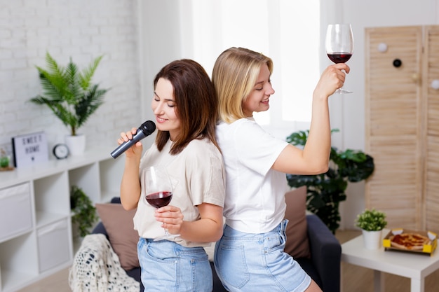 Domowa impreza - portret dwóch pięknych dziewczyn bawiących się razem, pijących wino, tańczących i śpiewających karaoke w przytulnym salonie