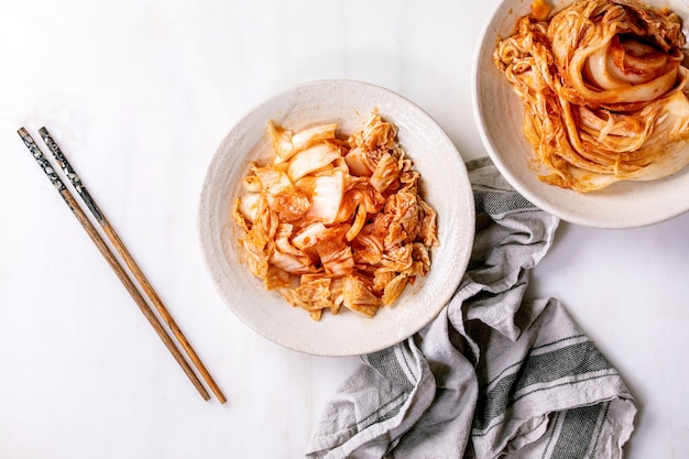 Domowa koreańska tradycyjna sfermentowana przekąska z kapusty kimchi w całości i posiekana, podawana na talerzu ceramicznym z pałeczkami na białej marmurowej ścianie. Leżał na płasko, przestrzeń