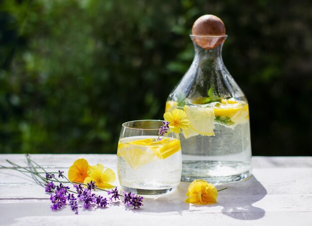 Domowa lemoniada z cytryną i kostkami lodu w szklanym dzbanku na białym drewnianym stole z żółtymi kwiatami i lawendą